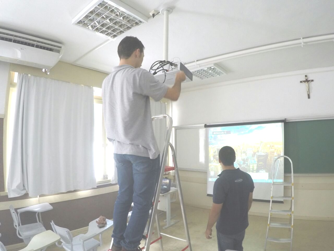 Sala de aula. Homem sobre uma escada conserta projetor. Outro homem está de pé a frente da projeção na tela